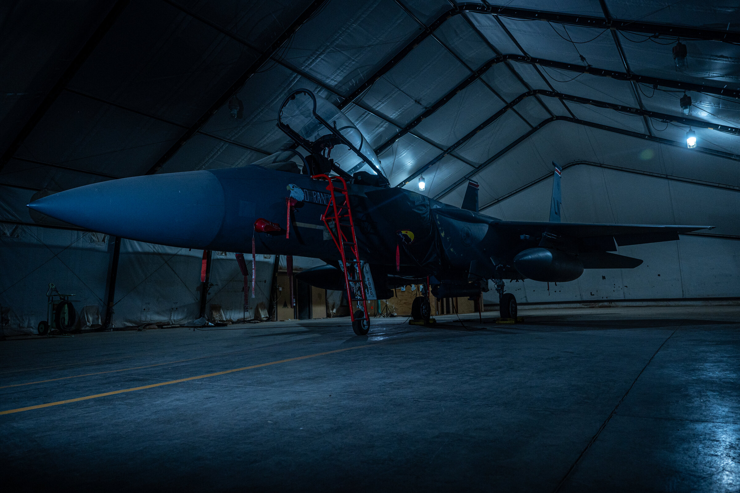 Dark hangar interior with a military fighter jet parked under bright overhead lights, red maintenance ladder positioned at the cockpit.