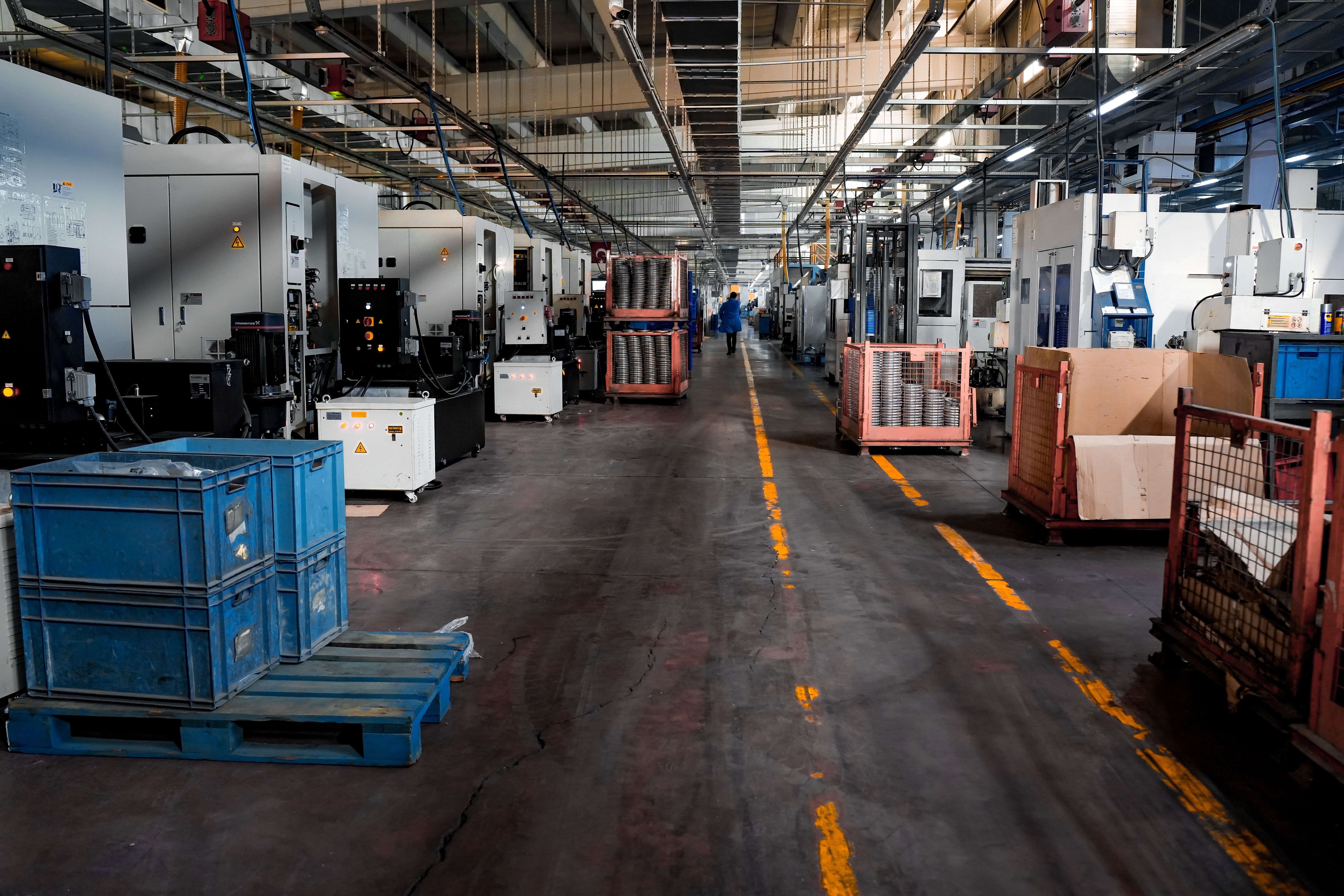 Manufacturing floor with rows of industrial CNC machines lining a central aisle under overhead pipes and fluorescent lighting.