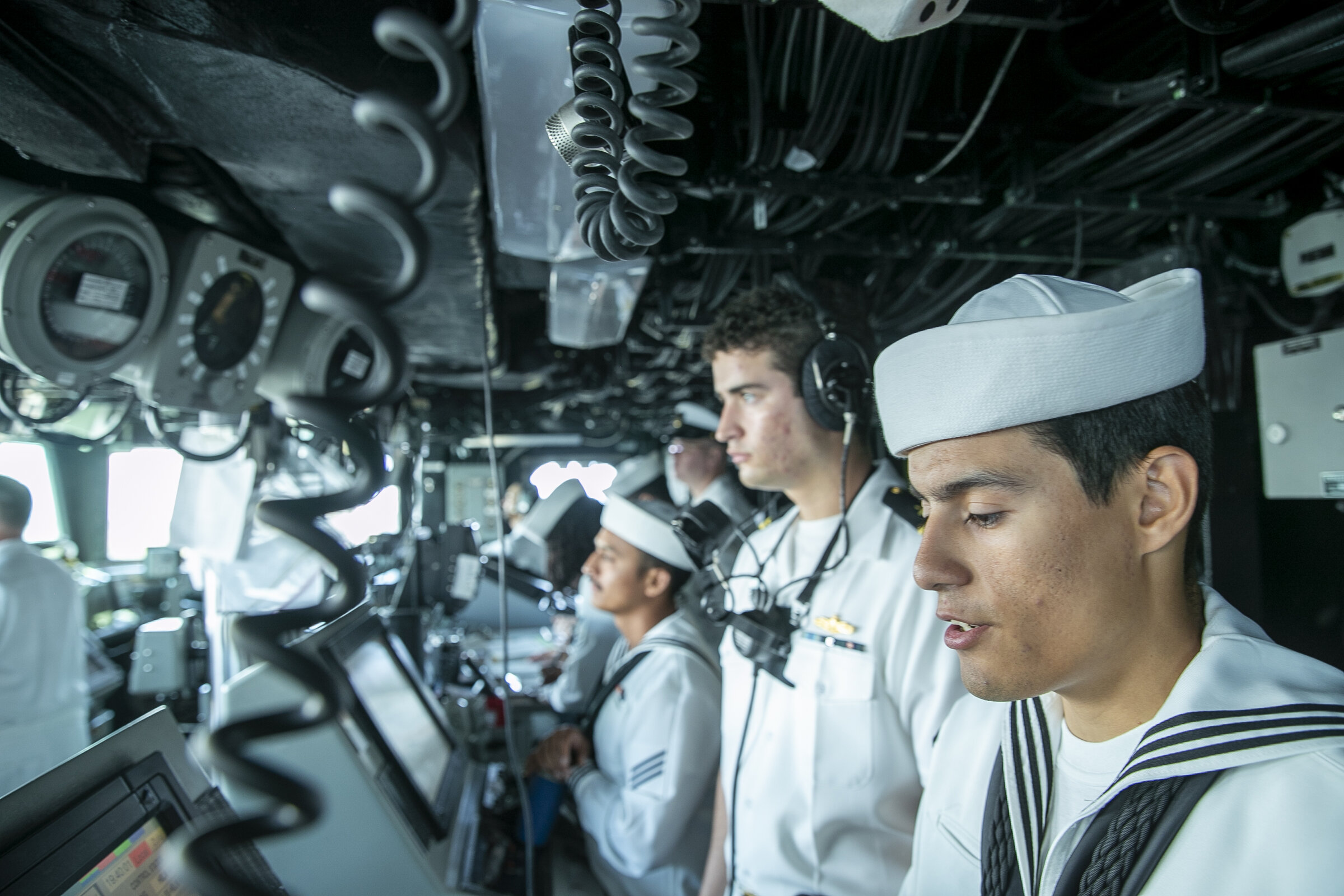 Sailors in uniform monitoring instruments and screens inside a naval vessel control room under low operational lighting.